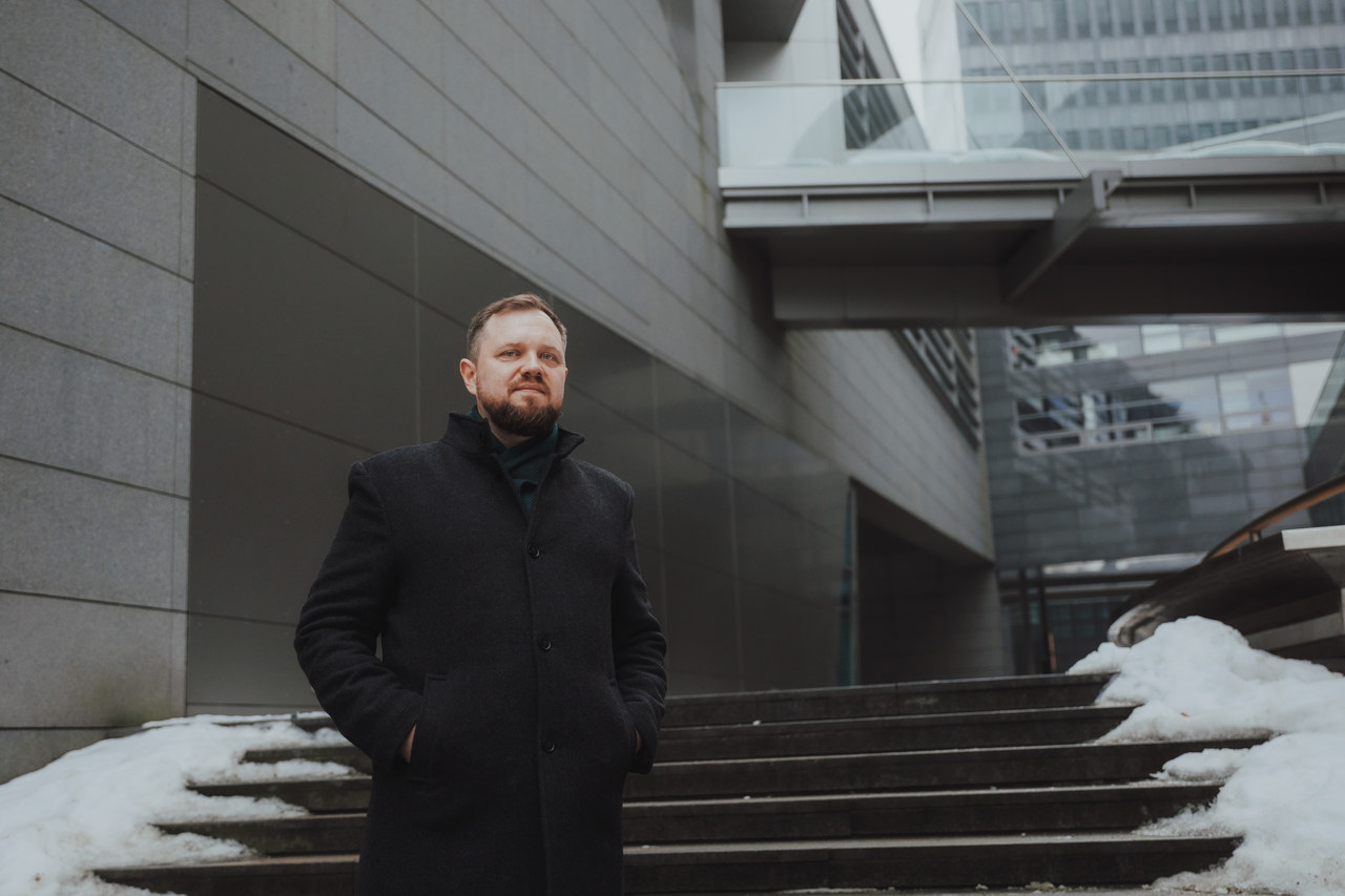 Łukasz Stocki w ciemnym wełnianym płaszczu na schodach przed nowoczesnym biurowcem zimą